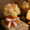 Bouquet de fleurs en cire vegetale dans une boite orange avec noeud en satin blanc,posé sur un livre décoratif.